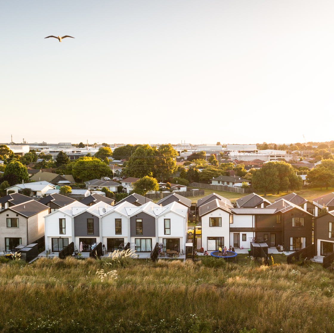 Home Capital Partners – row of modern townhouses with small backyards and a trampoline, viewed over a grassy reserve with the suburb in the background. Located in Broomfield, Christchurch.