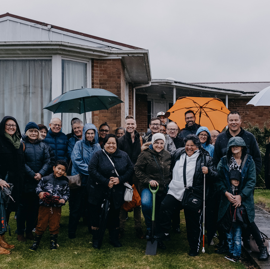 Home Capital Partners – group photo of community members outside a brick house on a rainy day, people with umbrellas and a ceremonial shovel.
