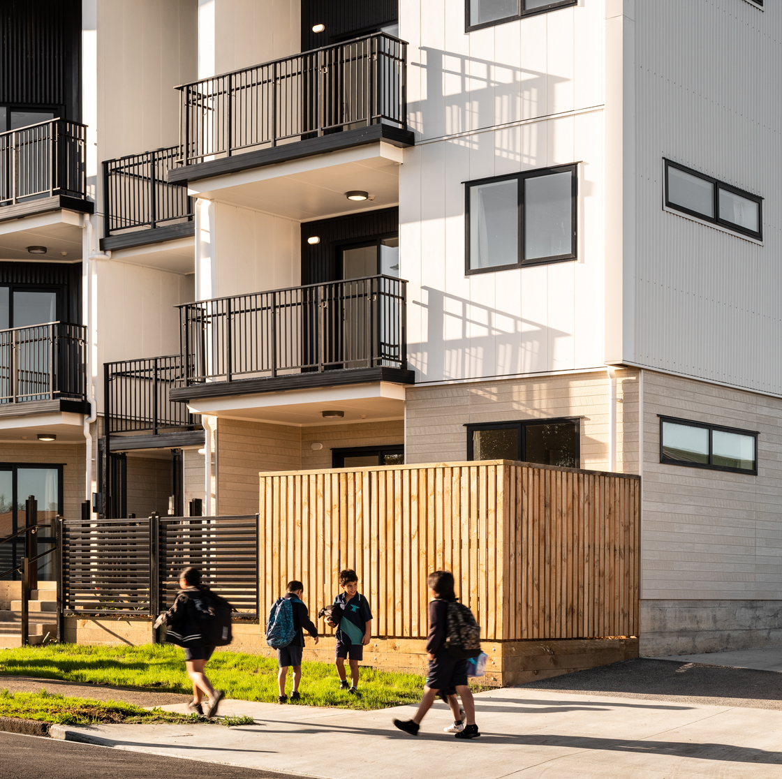 Home Capital Partners – exterior of a modern multi-storey apartment building located in Onehunga, Auckland, with black balconies; four schoolchildren with backpacks walking on the footpath in front.