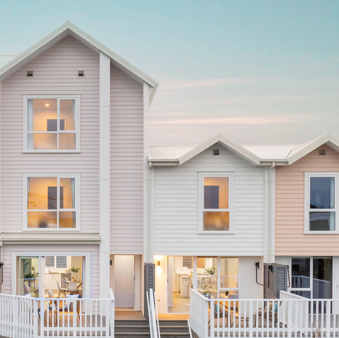 Home Capital Partners – row of modern pastel weatherboard townhouses with white railings and warm interior lights, located within the Te Pākau Maru development in New Brighton, Christchurch.