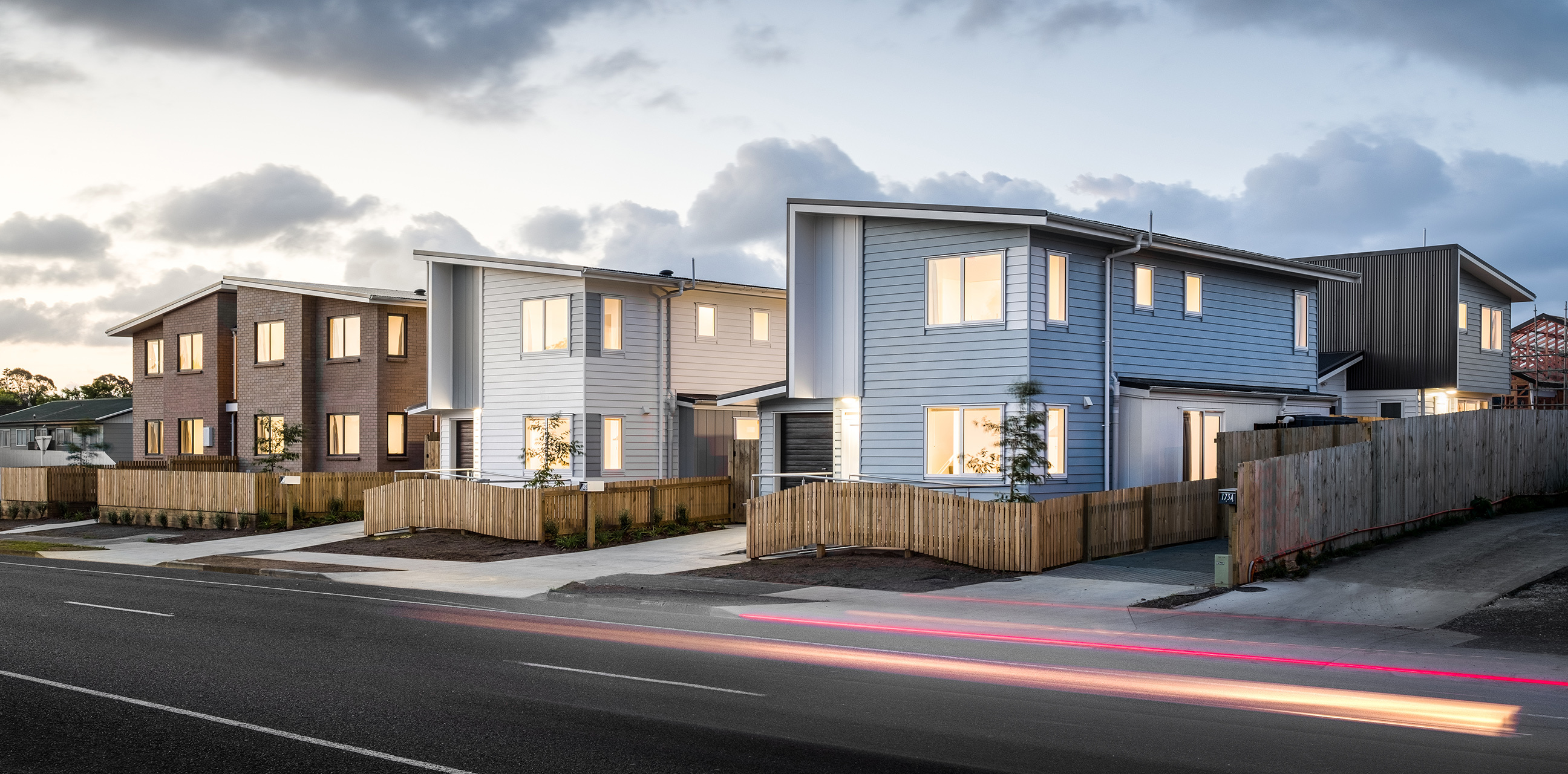 Home Capital Partners – street view of modern two-storey townhouses with timber fencing and lit windows; light trail from a passing car.